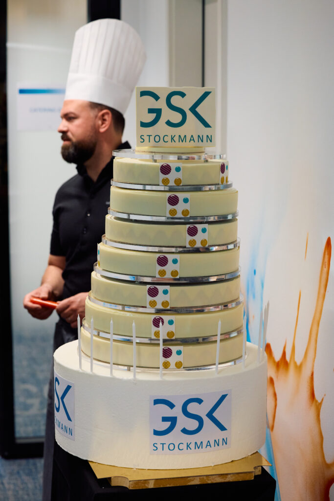 Chef in a tall hat standing behind a large multi-tier anniversary cake with the GSK Stockmann logo