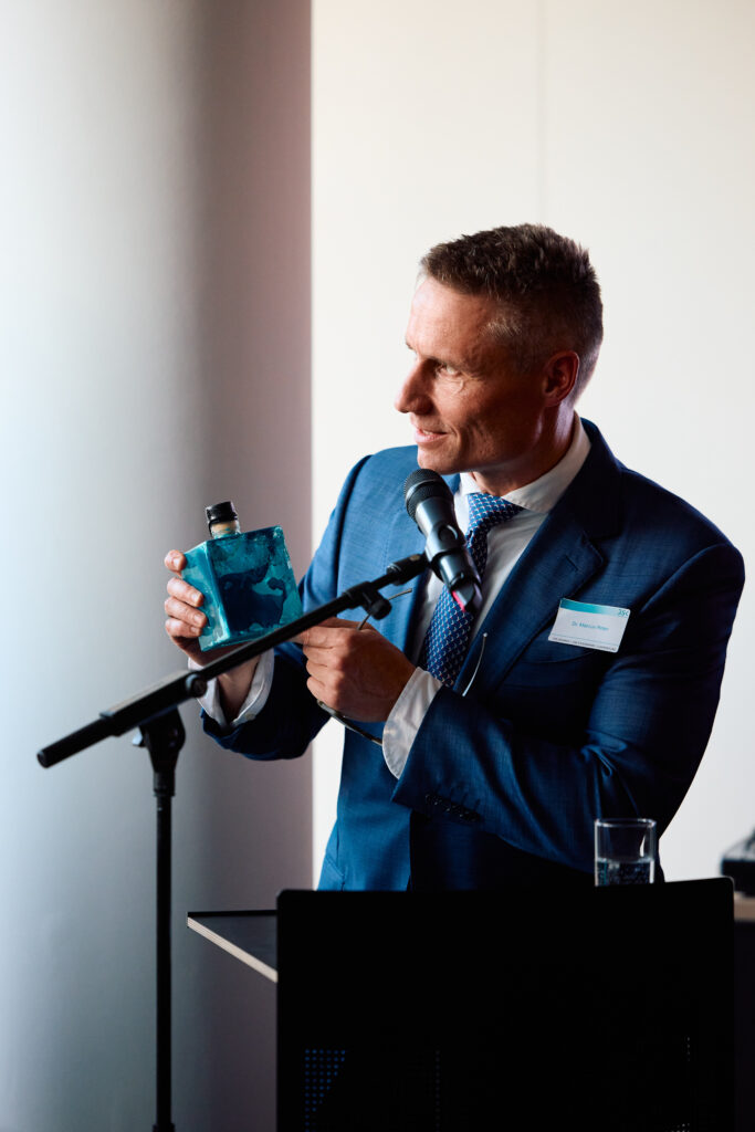 Man speaking at a lectern while holding a decorative bottle at the GSK Stockmann 10th anniversary event